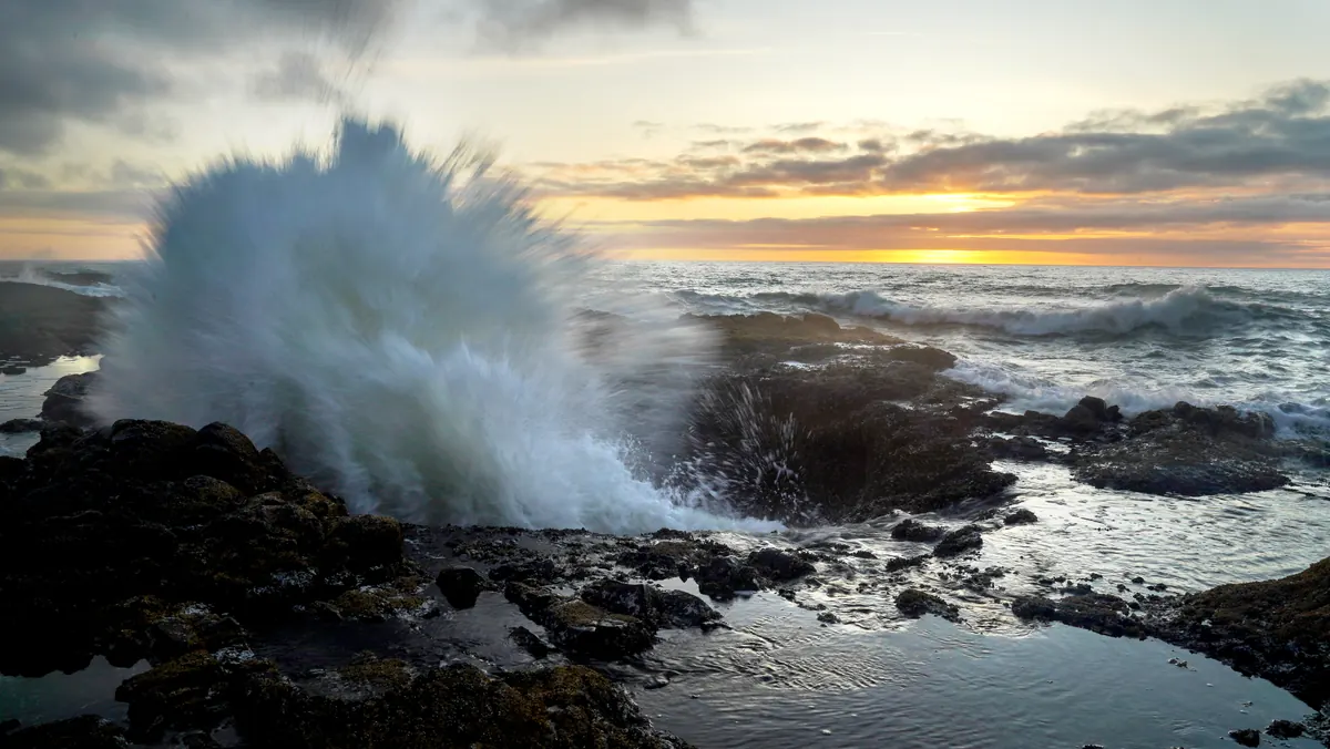 A wave bursts above the rocky shore at Thor’s Well, Cape Perpetua, under a warm sunset.